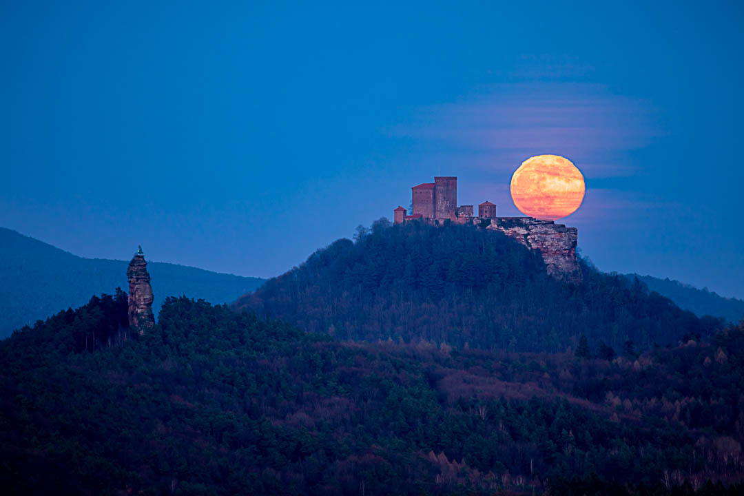 Vollmond über der Burg Trifels. Roter Vollmond verzerrt durch die Luftbewegungen zarte Wolkenschlieren, rechts noch der Kletterfelsen Asselstein