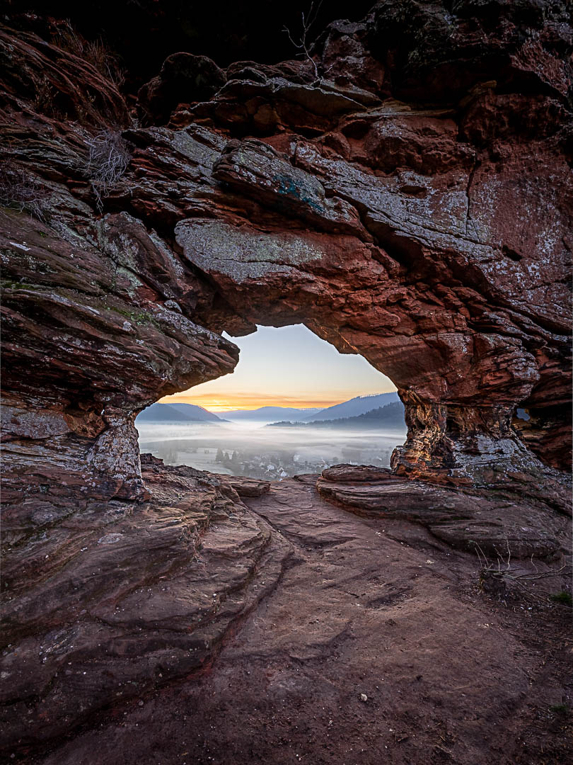 Das Felsentor am Wachtfels leuchtet im Morgenlicht und bietet einen Blick in ein nebelverhangenes Tal und der Morgenröte